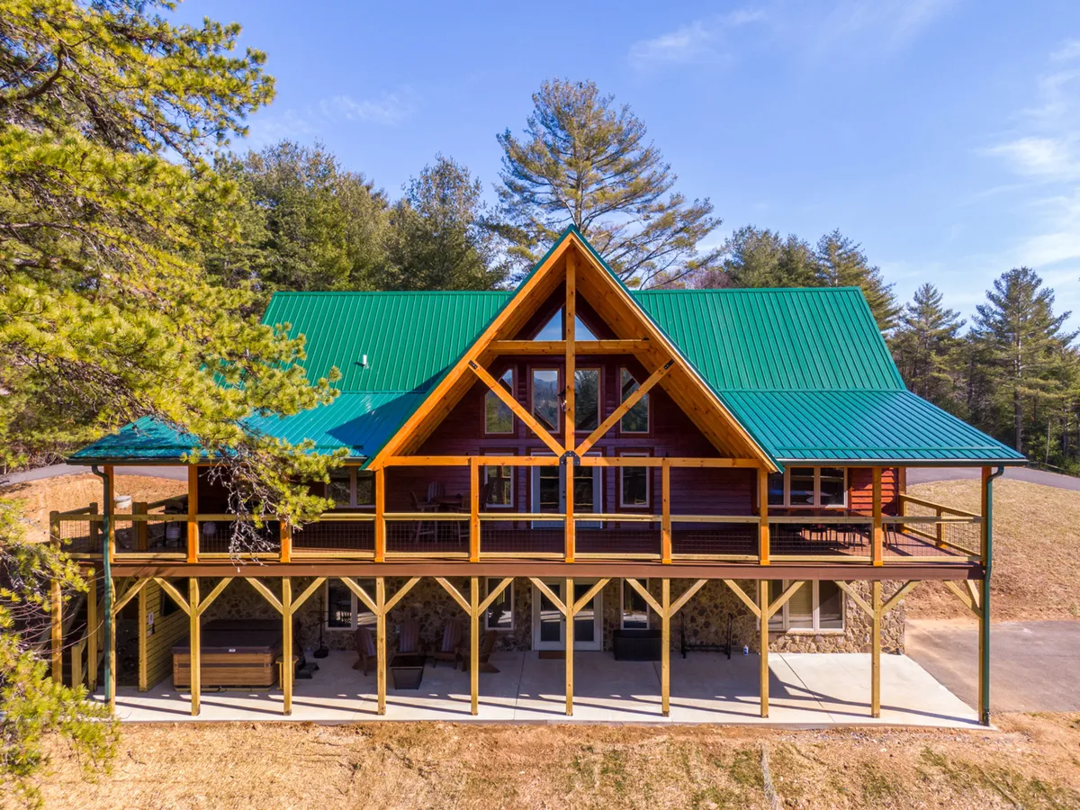 The image showcases the exterior of a beautiful single-family house with a striking green metal roof, expansive wooden deck, large windows that let in natural light, and a charming stone base. Set amidst lush greenery in a gated community, the house exudes a welcoming and serene atmosphere. The spacious deck is perfect for gathering with friends and family, enjoying barbecues, relaxing outdoors, and taking in tranquil views of the surrounding nature, with the promise of colorful sunsets and starry nights.
