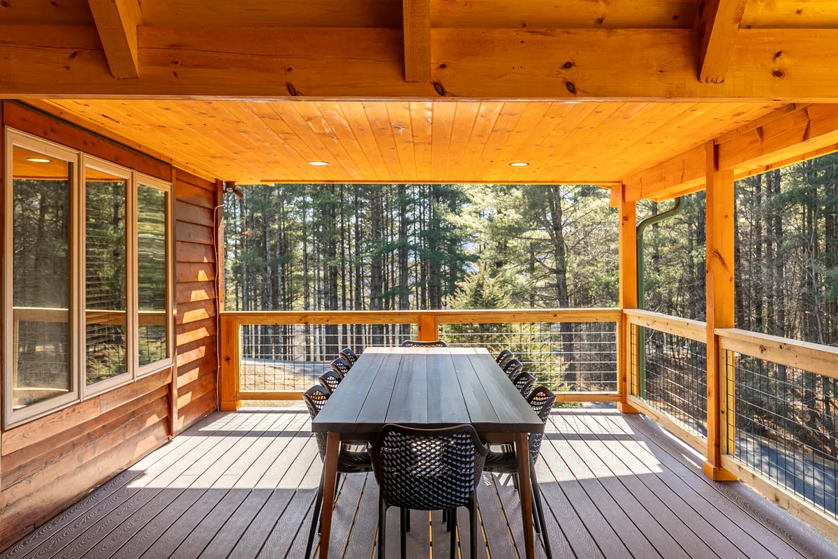 An outdoor dining area with a large wooden table set on a covered porch, surrounded by forest views. The setting suggests communal meals, morning coffee, or evening relaxation after a day of outdoor exploration, with nature always close at hand.