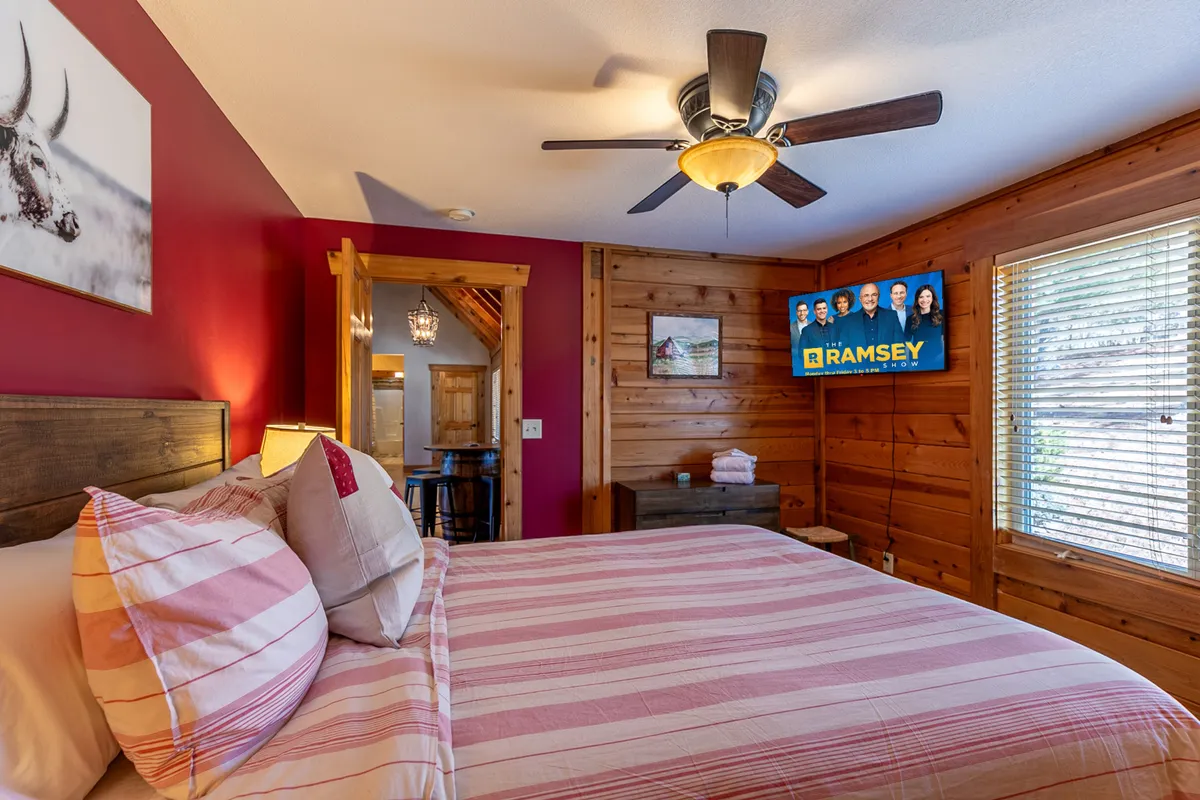 A cozy bedroom with a bed featuring striped bedding, wooden walls and ceiling, a mounted TV playing a show, and decorative art on the wall. The room feels intimate and inviting, representing the modern comforts paired with rustic charm.