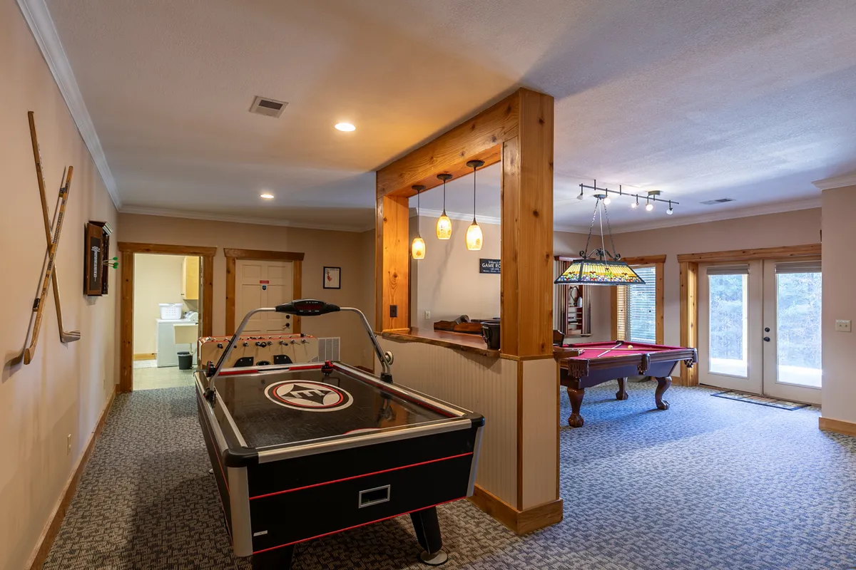 A well-lit recreational space featuring an air hockey table and pool table, with pendant lighting overhead and double doors leading to an outdoor area. The room feels spacious and energetic, emphasizing its role as a center for lively activities and gatherings.