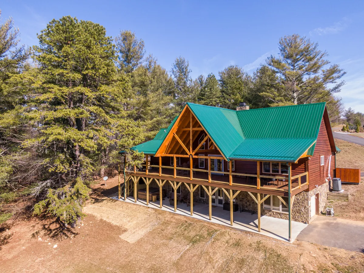 A rustic log cabin featuring a green metal roof, spacious front porch with sturdy wooden columns, and set amid lush greenery. The porch invites outdoor gatherings and relaxation, while the overall scene exudes a cozy, welcoming vibe ideal for family get-togethers.