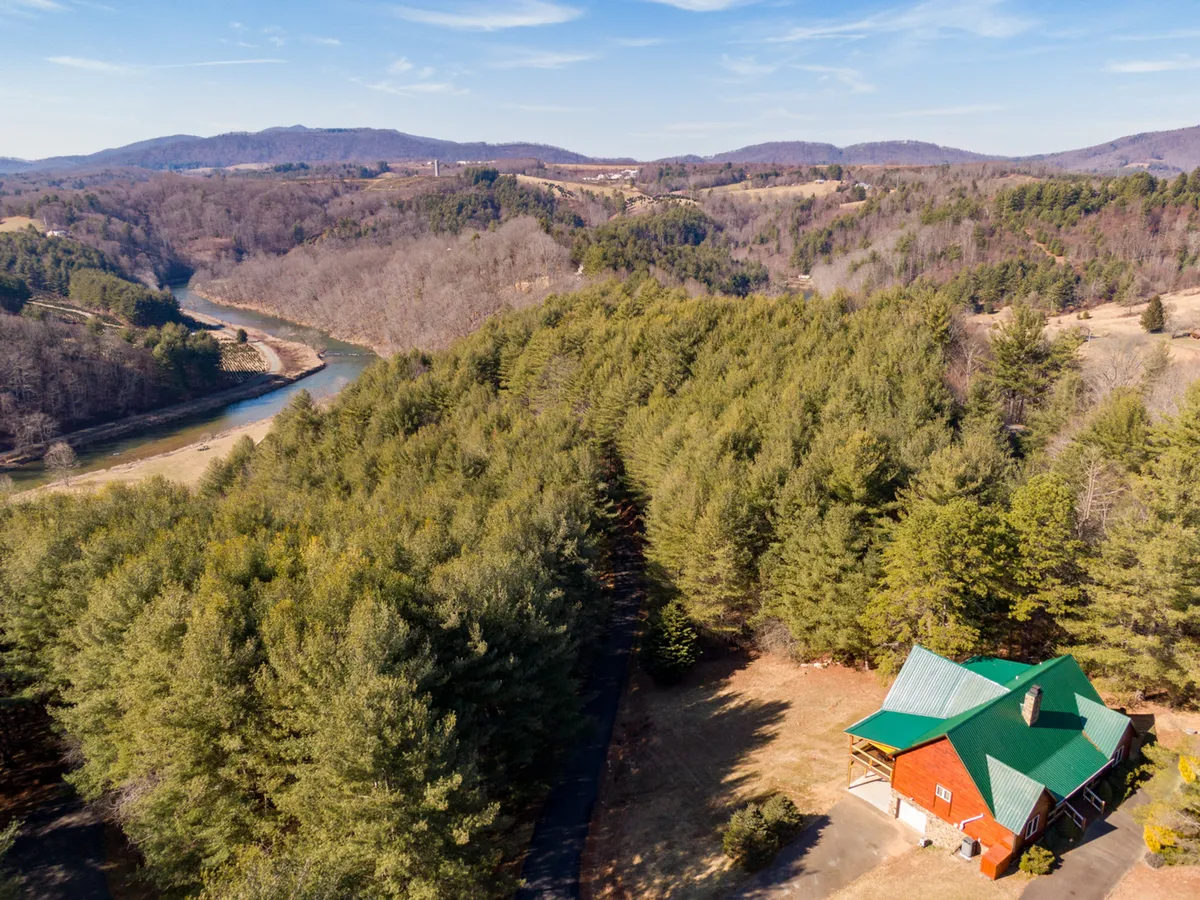 This image captures a rustic cabin nestled beside a winding river, with lush pine trees and distant mountains in the background. The river is prominent, suggesting opportunities for fishing, kayaking, or simply enjoying the tranquil waterside environment. The inviting setting hints at easy access to outdoor activities and serene nature immersion.