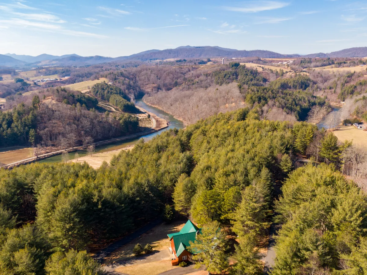 A serene view of a log cabin with a green roof situated near a meandering river, framed by tall trees and a dramatic mountain backdrop. The image radiates a quiet, untouched wilderness, inviting exploration and relaxation by the water.