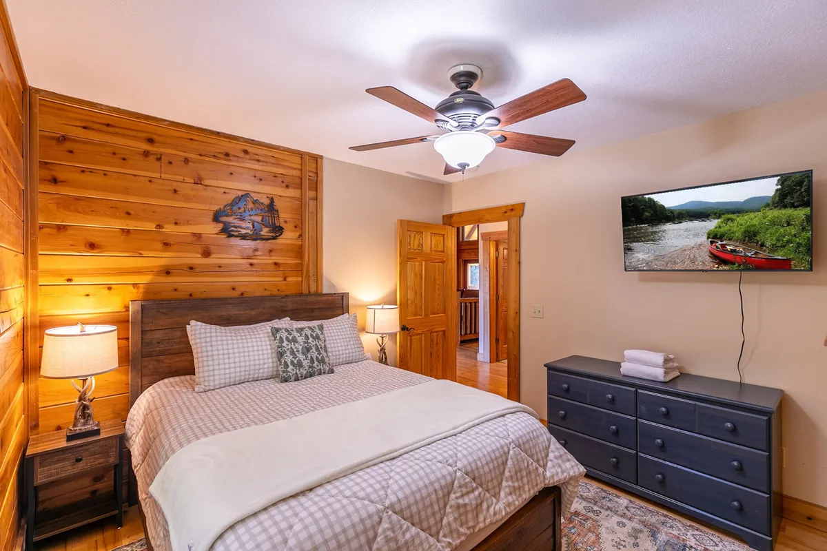 A tranquil bedroom featuring wooden wall paneling, a bed with decorative pillows, and a mounted TV displaying a scenic view. A nightstand with a lamp adds to the sense of coziness and comfort, emphasizing the guest-centric touches like quality linens and entertainment.