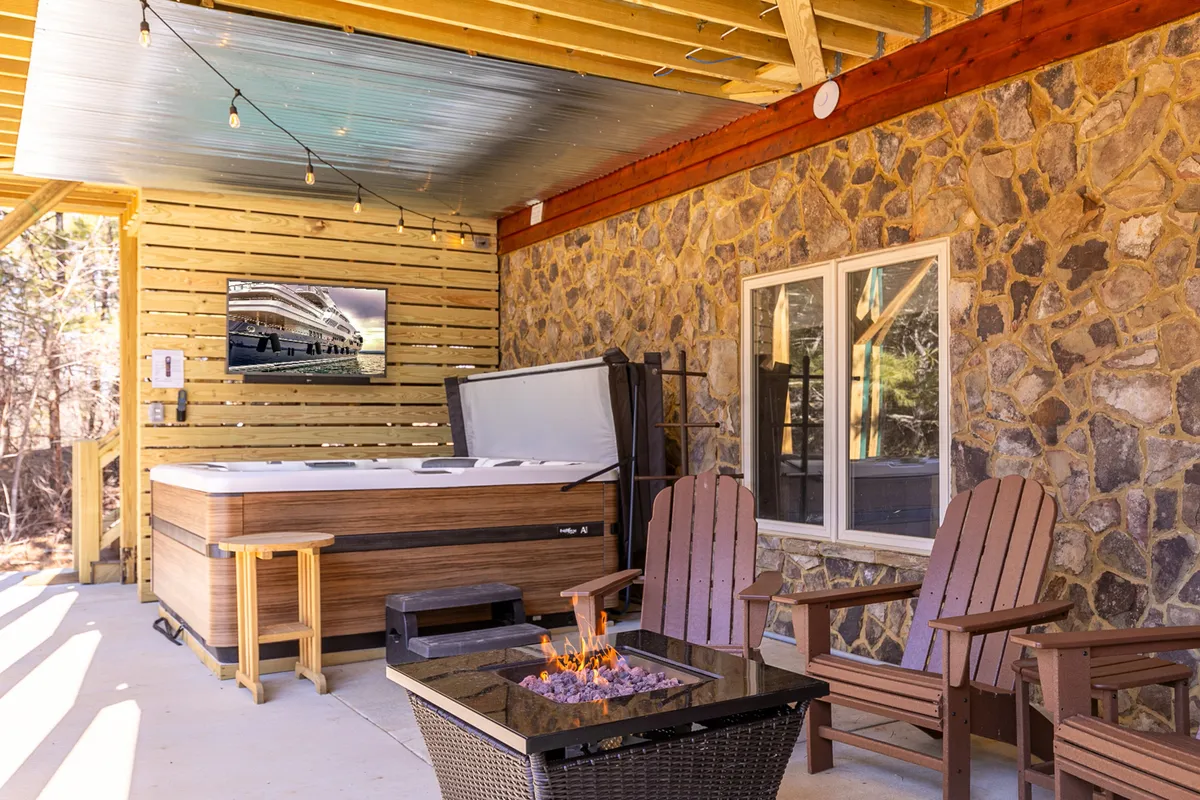 A lower-level patio area equipped with a large hot tub, a stone fire pit, wooden chairs, and an outdoor TV mounted on a stone wall. The string lights above add a warm, inviting glow, making it an ideal spot for relaxation and evening entertainment.