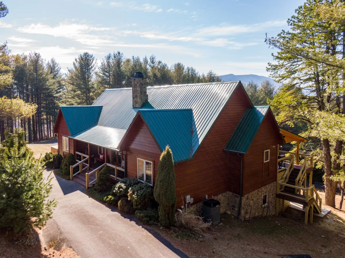 An inviting view of a wooden cabin with a blue metal roof nestled among trees, featuring a cozy porch area and a prominent stone chimney. The natural setting suggests easy access to outdoor adventures and trails.