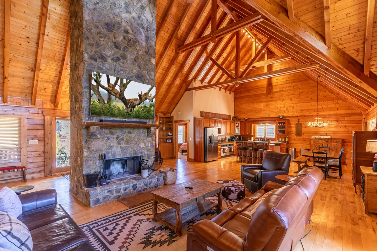 A spacious, open-concept living area showcasing a magnificent stone fireplace as the focal point. High wooden ceilings and large windows fill the space with natural light. Comfortable seating is arranged for group gatherings, while a rustic dining table and nearby kitchen area invite both conversation and shared meals.